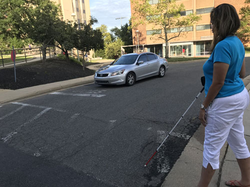 picture shows a student with a white cane standing at the crosswalk with a stop sign next to it on the students's left,  There is a vehicle approaching from her right from about 10 feet away.